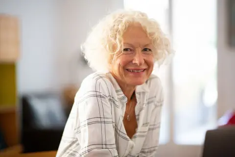 Une femme souriante aux cheveux bouclés blonds, portant une chemise à carreaux. Son état civil célibataire rayonne de joie.
