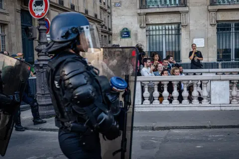 Policjant w pełnym rynsztunku ochronnym z tarczą, obserwuje tłum za barierką.