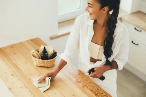 Mujer sonriente limpiando una mesa de madera sin tratar, usando un paño y spray para quitar manchas de aceite.