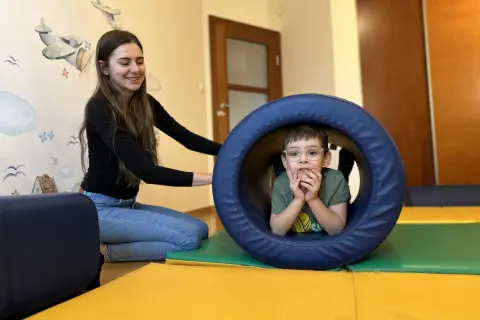 Chłopiec w okularach w tunelu terapeutycznym, obok uśmiechnięta terapeutka.