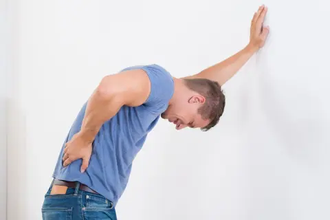 Uomo con forte mal di schiena da freddo, cerca rimedi. Si appoggia al muro con una mano, piegato dal dolore.