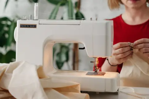 Biała maszyna do szycia na metalowych podzespołach, z nawiniętą białą nicią, gotowa do pracy. Kobieta w czerwonej bluzce przygotowuje materiał.