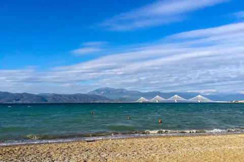 Spokojne morze i plaża w Grecji w styczniu. W oddali widać most i góry.