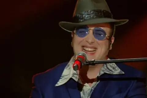 Un artista con cappello e occhiali da sole canta al microfono, un talento tra i cantanti marchigiani.