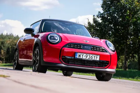 Czerwony Mini Cooper na tle zieleni. Wiele osób szuka opinii o tym modelu, a ten egzemplarz wygląda na gotowy do jazdy.