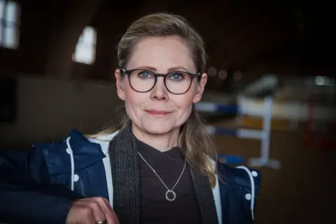 Anna Schreil med glasögon och en blå jacka, står i en ridhall.