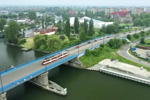 Tram z czerwonymi pasami jedzie przez most nad rzeką, w tle widać miasto i tereny przemysłowe, jak na plaży Stogi.