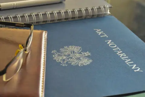 Na niebieskiej okładce widnieje napis "AKT NOTARIALNY" i orzeł. Obok leżą okulary i notatnik.
