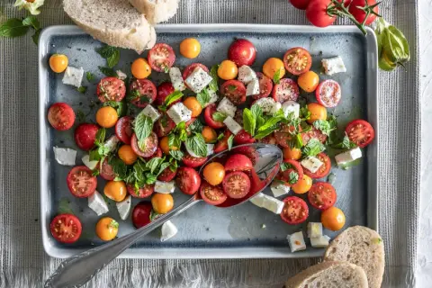 Frischer Tomate Mozzarella Salat mit bunten Kirschtomaten, Feta und Minze, serviert auf einem grauen Teller mit Brot.
