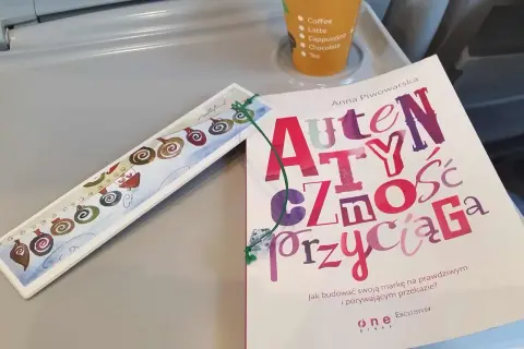 Książka "Autentyczność przyciąga" Anny Piwowarskiej na stoliku, obok ozdobna zakładka i kubek z napojami.