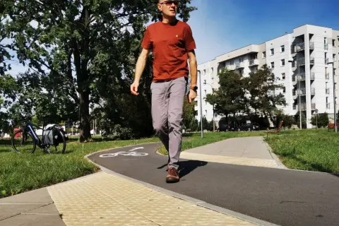 Mężczyzna idzie ścieżką rowerową, na której widnieje symbol roweru. Obok ścieżki znajduje się chodnik z wypukłą nawierzchnią, sugerujący przejście dla pieszych.