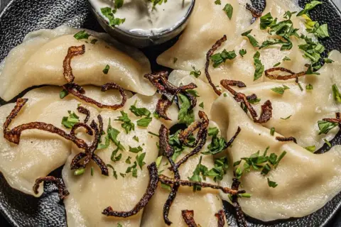 Pyszne pierogi z ziemniakami i boczkiem, posypane chrupiącą cebulką i natką pietruszki. Podane z kleksem śmietany.
