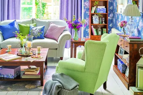 Jasny salon z fioletowymi zasłonami i kwiecistą tapetą. Wygodna kanapa i zielony fotel tworzą przytulną atmosferę, idealną na wiosnę w domu.