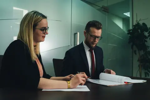 Kobieta i mężczyzna w okularach siedzą przy biurku i przeglądają dokumenty.