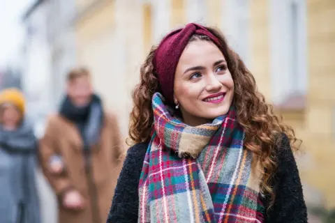 Uśmiechnięta dziewczyna w bordowej opasce i kolorowym szalu, idealnym do płaszcza w kratę. W tle para spacerująca ulicą.