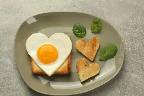 Jajko sadzone w kształcie serca na grzance, obok tostów w kształcie serc i liści szpinaku.