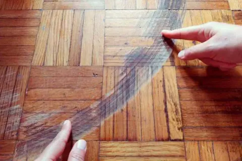 Manos señalan una mancha oscura en un suelo de parquet de madera. Se muestra cómo disimular manchas en parquet.