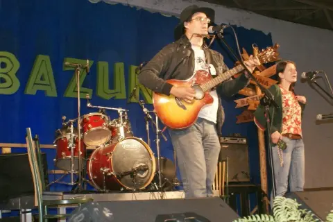 Rafał Chojnacki gra na gitarze i harmonijce ustnej, obok niego śpiewa kobieta. W tle zespół muzyczny i niebieska kurtyna z napisem "BAZUNA".