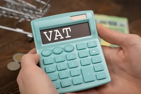 Kalkulator z napisem VAT w rękach, symbolizujący zwolnienie podmiotowe z VAT. W tle monety i banknoty.