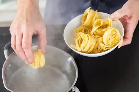 Ręce wkładają porcję makaronu do gotującej się wody. W misce obok czeka kolejna porcja, by uzyskać idealny makaron al dente.