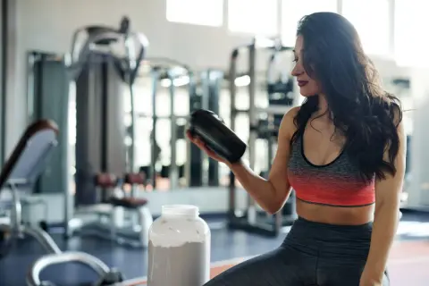Frau im Fitnessstudio hält einen Shaker, daneben ein großer Behälter mit Pulver. Perfekt für den **proteinshake vor oder nach dem training**.