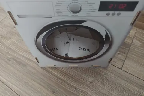 Kartonowa pralka z bębnem pełnym kart z napisami "GAZETA", "ZKA", "ARBA".