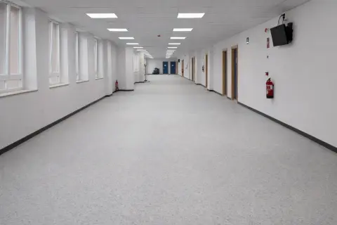 Pasillo largo y luminoso de un colegio, con ventanas a la izquierda y puertas a la derecha. El suelo del colegio es de un material gris moteado.