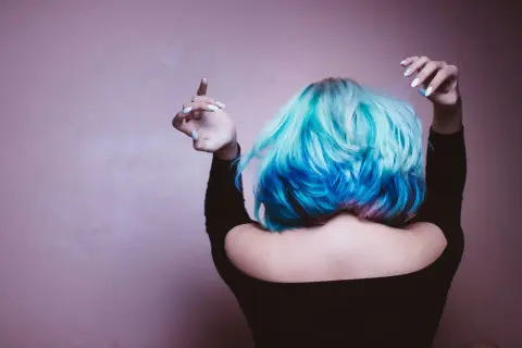 Dziewczyna z turkusowo-niebieską peruką kok, z różowymi akcentami, w czarnej bluzce z odkrytymi ramionami, unosi ręce.