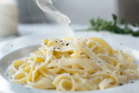 En krämig portion pasta alfredo med ånga som stiger, toppad med riven parmesan och svartpeppar.
