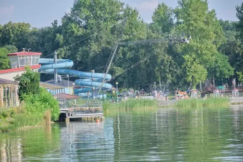 Letnia zabawa na **zalewie zemborzyckim**: niebieskie zjeżdżalnie wodne, ludzie kąpiący się i relaksujący się na plaży, otoczeni zielenią.