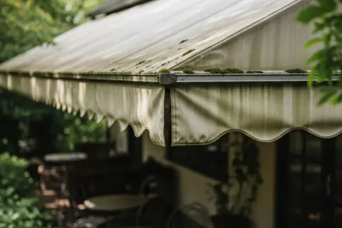 Tenda da sole con muschio e segni di lavaggio. L'esterno di un locale con tavoli e sedie visibili sotto.