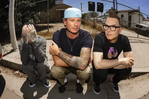 Die Mitglieder von blink-182 hocken auf dem Bürgersteig. Links Travis Barker, der sein Gesicht verdeckt. In der Mitte Mark Hoppus mit Cap und tätowierten Armen. Rechts Tom DeLonge mit Brille und Irokesenschnitt.
