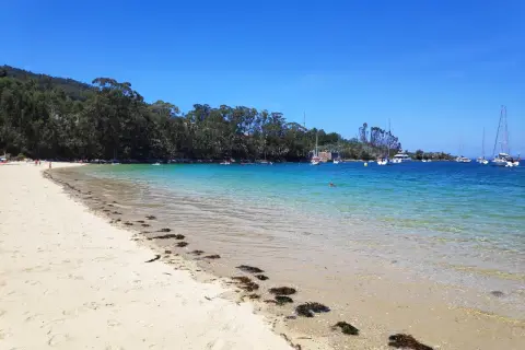 Arena dorada y aguas cristalinas en la playa de Arneles, con veleros anclados y un cielo azul intenso.