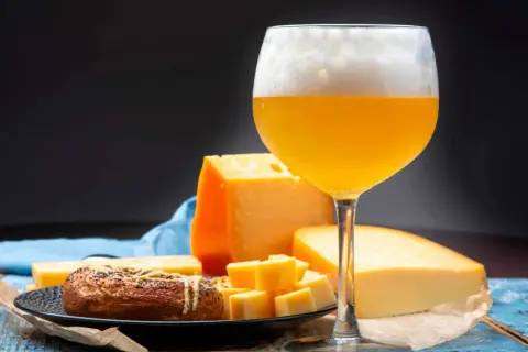 Un bicchiere di birra dorata con schiuma abbondante, accompagnato da vari tipi di formaggio e un panino con semi.