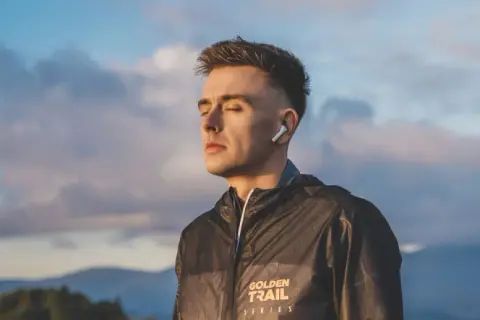 Młody mężczyzna w słuchawkach ANC, z zamkniętymi oczami, w kurtce z napisem "GOLDEN TRAIL SERIES", na tle nieba i gór.