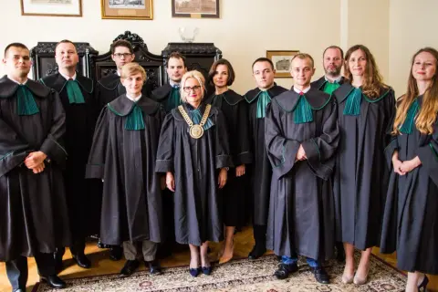 Grupa prawników w togach stoi w pomieszczeniu z meblami i obrazami.