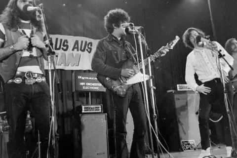 Band auf der Bühne, Gitarrist mit Afro, Sänger mit Schnurrbart, 70er Jahre Rock.