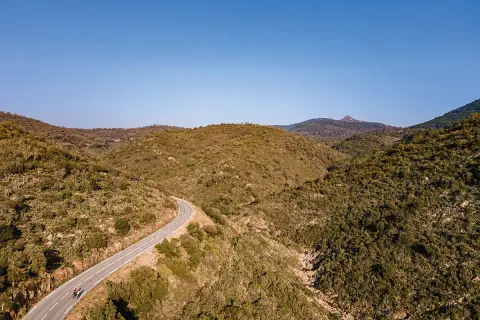 Vue aérienne d'une route sinueuse traversant des collines verdoyantes, menant vers le col du Testanier. Deux cyclistes s'engagent sur la pente.