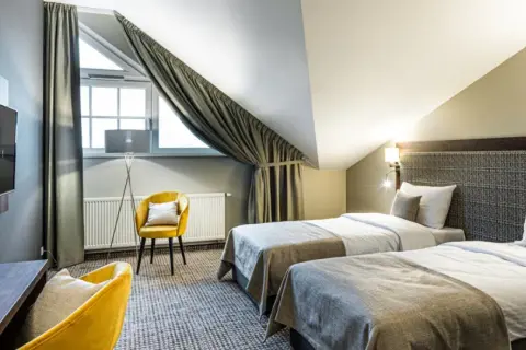 Pokój hotelowy z dwoma łóżkami, żółtym fotelem i biurkiem. Idealne miejsce na nocleg, gdzie gwiazdki w hotelach są gwarancją komfortu.