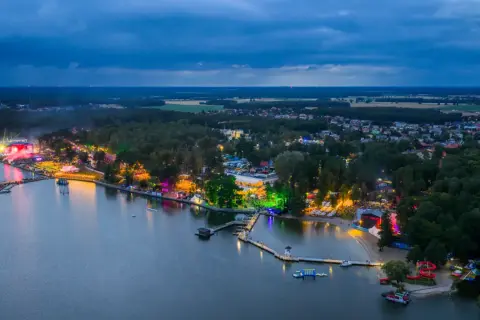 Nocny widok na jezioro i rozświetlone tereny imprezowe. Widać scenę, tłumy ludzi i kolorowe światła, które tworzą atmosferę sława festiwal.