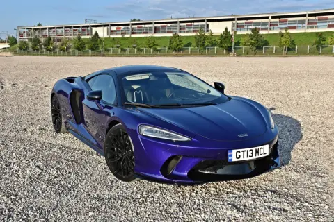 Elegancki, fioletowy McLaren GT na żwirowym placu, z nowoczesnym budynkiem w tle.