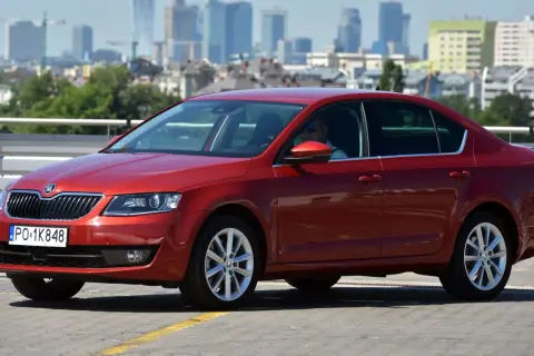 Czerwona Skoda Octavia 3 z widocznymi żarówkami w przednich reflektorach, zaparkowana na tle miasta.