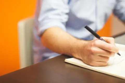 Mężczyzna pisze odręcznie notatki w zeszycie, przygotowując się do napisania prośby o dofinansowanie. Obok leży laptop.