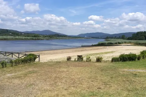 La playa fluvial Vilarello, con arena dorada y aguas tranquilas, se extiende ante colinas verdes bajo un cielo azul con nubes blancas.