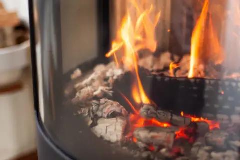 Glühende Kohlen und Flammen in einem modernen Kaminofen. Links daneben ein Korb mit Holzscheiten.