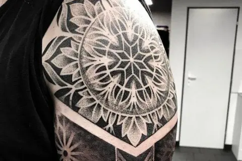 Imponujący mandala tatuaż na ramieniu, wykonany techniką dotwork. Wzór przypomina kwiat lub słońce, otoczony geometrycznymi kształtami.