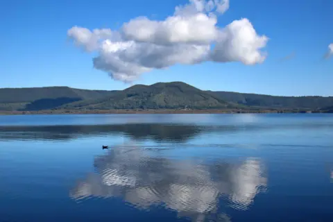 Un'oasi di pace al lago di Vici, con un cielo azzurro punteggiato da nuvole bianche che si specchiano sull'acqua calma.