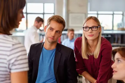 Ein Mann und eine Frau hören aufmerksam zu, während eine Kollegin spricht. Ein Beispiel für **talent mobility** in einem modernen Büro.