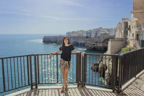 Donna sorridente con occhiali da sole si appoggia a una ringhiera con vista sul mare cristallino e le case bianche di Polignano a mare.