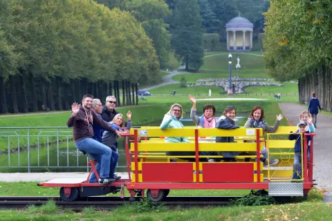 Fröhliche Menschen genießen eine Draisinenfahrt NRW durch eine Parklandschaft mit einem historischen Gebäude im Hintergrund.
